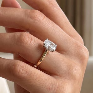Könnte beinhalten: Ein Goldring mit einem einzelnen rechteckigen Diamanten im Radiant-Schliff, der in einer Vier-Krappen-Fassung sitzt. Die Ringschiene ist glatt und poliert und zeigt ein klassisches Design, das die Reinheit und Brillanz des Steins betont.