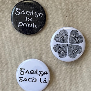 Puede incluir: Tres botones redondos con texto. Un botón negro tiene las palabras "Gaeilge is punk" en blanco. Un botón blanco tiene las palabras "Gaeilge sach lá" en negro. Un botón blanco tiene un diseño de trébol de cuatro hojas con las palabras "thú, do, mo, chuile" en negro.