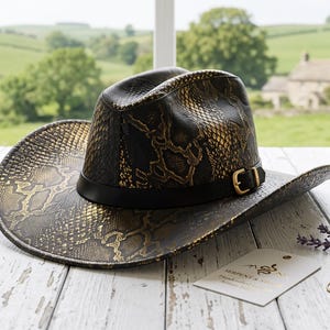 May include: A cowboy hat featuring a black and gold snakeskin pattern with a black leather band and a gold-toned metal buckle. A small white rectangular tag with the text Serpent & Soul is placed next to a brass compass and sprigs of lavender.