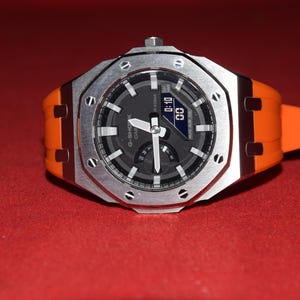 May include: A silver and black wristwatch with an orange rubber strap. The watch face has a black background with white markings and a digital display that reads "01:00".