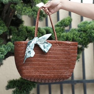 Puede incluir: Un bolso tote tejido marr&oacute;n con asas trenzadas y un pa&ntilde;uelo decorativo atado en la parte delantera. El bolso tiene forma redondeada y lo sostiene una mano. El pa&ntilde;uelo tiene un fondo blanco con un estampado floral verde.