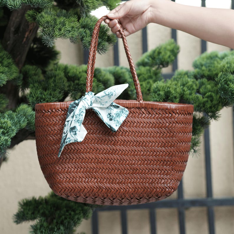 Puede incluir: Un bolso tote tejido marr&oacute;n con asas trenzadas y un pa&ntilde;uelo decorativo atado en la parte delantera. El bolso tiene forma redondeada y lo sostiene una mano. El pa&ntilde;uelo tiene un fondo blanco con un estampado floral verde.