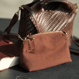 Pu&ograve; includere: Una pochette in tela marrone con cerniera dorata e un'etichetta in pelle con la scritta "Serena". Sullo sfondo una borsa a mano in pelle marrone intrecciata. Un taccuino in pelle marrone si trova nell'angolo in basso a sinistra. Le borse sono su una superficie scura.