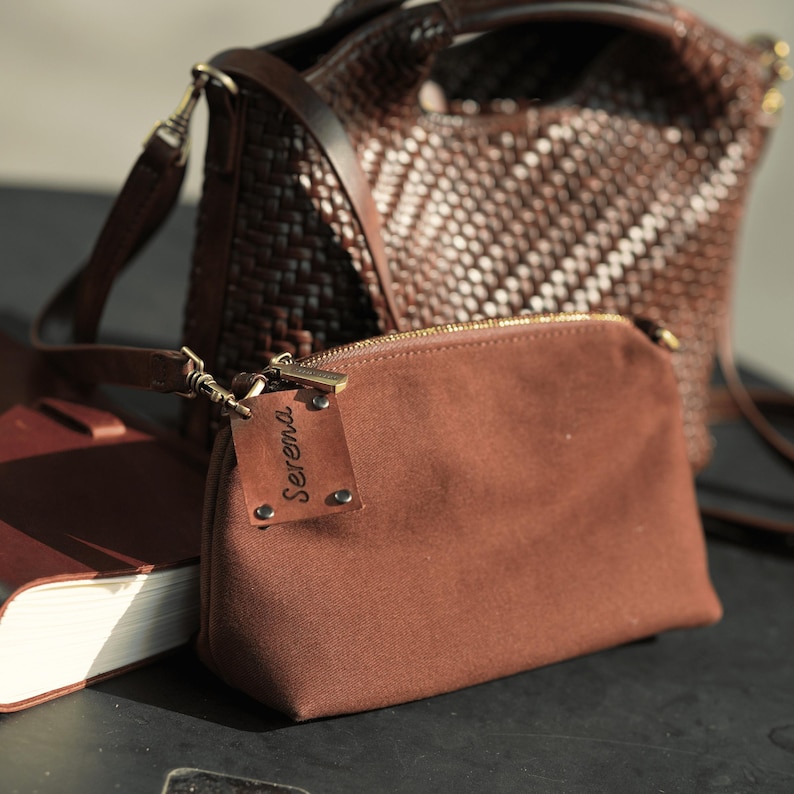 Pu&ograve; includere: Una pochette in tela marrone con cerniera dorata e un'etichetta in pelle con la scritta "Serena". Sullo sfondo una borsa a mano in pelle marrone intrecciata. Un taccuino in pelle marrone si trova nell'angolo in basso a sinistra. Le borse sono su una superficie scura.