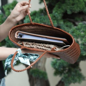 Puede incluir: Un bolso tote marr&oacute;n tejido con asas trenzadas. El bolso est&aacute; abierto, revelando una tableta, un cuaderno y una bolsa con cord&oacute;n. Una bufanda estampada est&aacute; atada a una de las asas. El bolso se sostiene contra un fondo de follaje verde.
