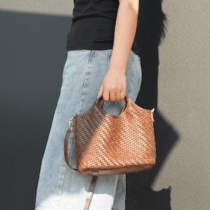 Pu&ograve; includere: Una borsa a mano in pelle intrecciata marrone con un manico superiore e una tracolla staccabile. La borsa ha una base rettangolare e un motivo intrecciato. La borsa &egrave; tenuta da una persona che indossa una maglietta nera e jeans blu.