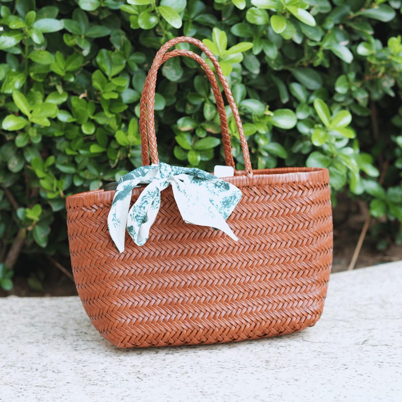 Puede incluir: Un bolso tote tejido marr&oacute;n con asas trenzadas. Una bufanda estampada en blanco y verde est&aacute; atada a la bolsa. La bolsa est&aacute; hecha de un material tejido y est&aacute; sobre una superficie de hormig&oacute;n.