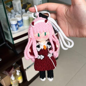 May include: A crochet doll keychain featuring a character with long pink hair, two small red horns, and green eyes. The doll wears a dark red dress with white accents, a small orange detail at the collar, and black trousers with white shoes, attached to a white lanyard.