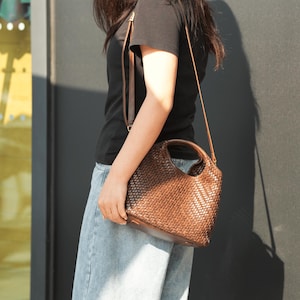 Pu&ograve; includere: Una borsa a mano in pelle intrecciata marrone con un manico superiore e una tracolla staccabile. La borsa ha una forma arrotondata e un design strutturato a trama di vimini. La borsa &egrave; portata da una persona che indossa una maglietta nera e jeans blu.