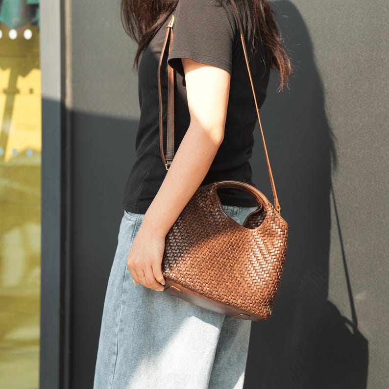 Pu&ograve; includere: Una borsa a mano in pelle intrecciata marrone con un manico superiore e una tracolla staccabile. La borsa ha una forma arrotondata e un design strutturato a trama di vimini. La borsa &egrave; portata da una persona che indossa una maglietta nera e jeans blu.