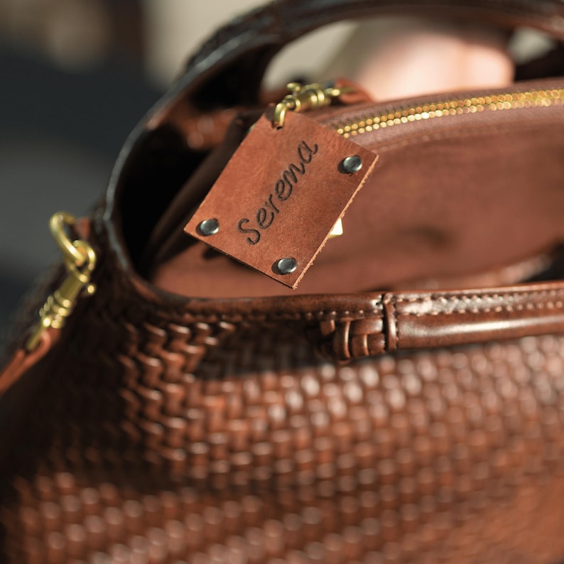 Pu&ograve; includere: Primo piano di una borsa in pelle marrone con una trama intrecciata. La borsa ha un manico superiore e una cerniera dorata. Un piccolo cartellino rettangolare in pelle con il nome "Serena" &egrave; attaccato.