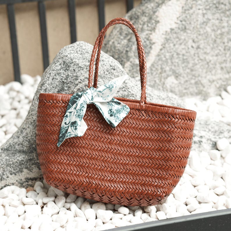 Puede incluir: Un bolso tote tejido marr&oacute;n con asas trenzadas y una bufanda decorativa. El bolso est&aacute; hecho de un material tejido apretado, y la bufanda tiene un fondo blanco con un estampado floral verde. El bolso est&aacute; sobre guijarros blancos.