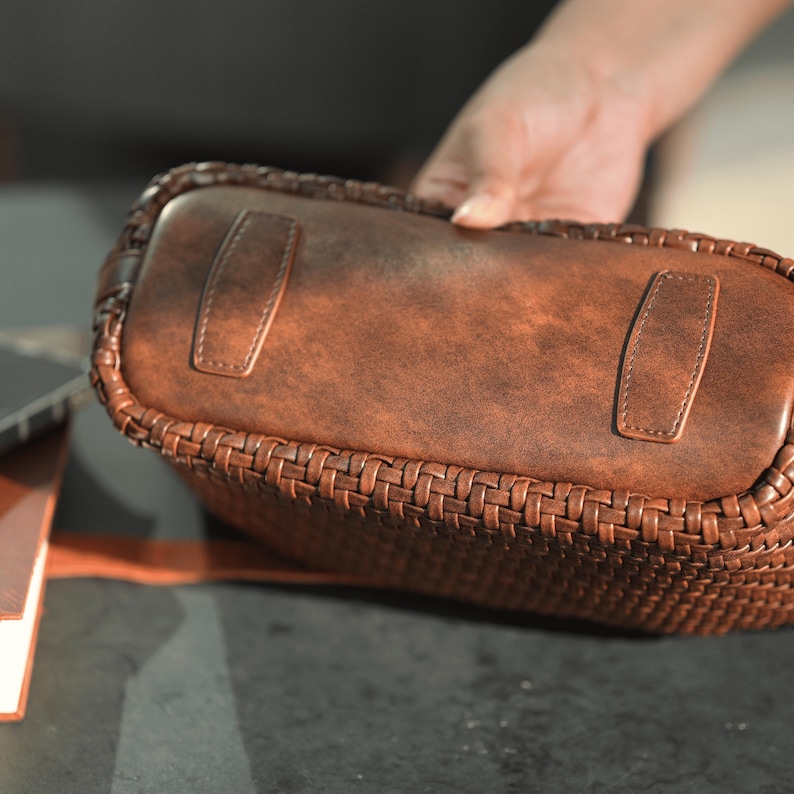 Pu&ograve; includere: Una borsa in pelle marrone con un design intrecciato sui lati e una base in pelle liscia. La borsa ha due inserti rettangolari in pelle sulla base. La borsa &egrave; tenuta da una mano.