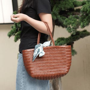 Puede incluir: Un bolso tote tejido marr&oacute;n con asas trenzadas. El bolso tiene forma redondeada y est&aacute; decorado con una bufanda estampada atada a una de las asas. El bolso est&aacute; hecho de un material tejido.