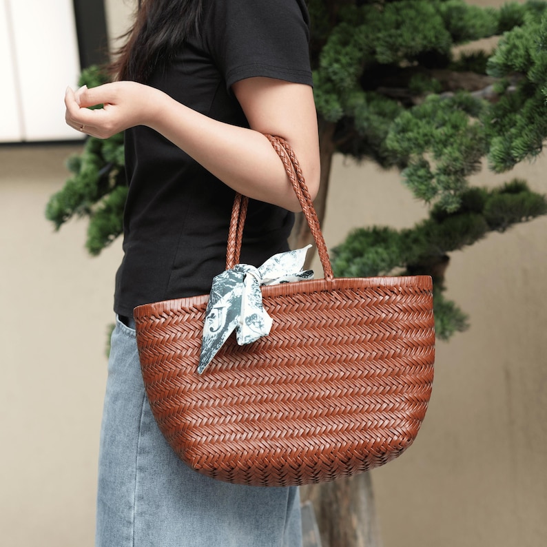 Puede incluir: Un bolso tote tejido marr&oacute;n con asas trenzadas. El bolso tiene forma redondeada y est&aacute; decorado con una bufanda estampada atada a una de las asas. El bolso est&aacute; hecho de un material tejido.