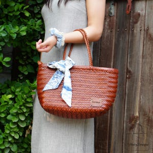 Puede incluir: Un bolso tote de cuero marr&oacute;n tejido con asas trenzadas. Una bufanda con estampado azul y blanco est&aacute; atada al asa. La bolsa tiene una peque&ntilde;a etiqueta de cuero que dice "Serena".