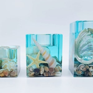 May include: Three teal blue glass candle holders with a square shape. Each holder is filled with a clear resin and contains various seashells and starfish. The largest holder features a large abalone shell.