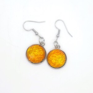 May include: A pair of dangle earrings with a silver-colored metal frame. Each earring features a round, textured, orange-yellow glass or resin cabochon. The earrings have a simple hook closure.
