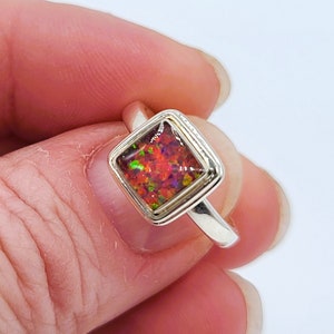 May include: A silver ring featuring a square-cut opal gemstone. The opal displays a vibrant array of colors, including red, orange, and green. The ring's band is a simple, polished silver.