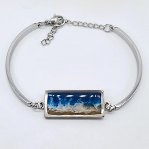 May include: A silver bangle bracelet with a rectangular resin charm featuring a blue and white ocean wave design.