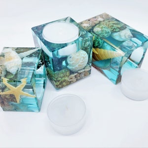 May include: Three teal blue glass candle holders with seashells and starfish embedded inside. Each holder has a small, clear glass votive candle inside.