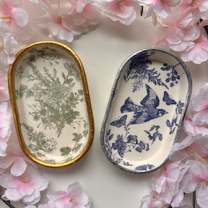 May include: Two decorative oval trays. One has a gold rim and a green floral design on a cream background. The other has a silver rim and a blue bird and butterfly design on a cream background. Both are surrounded by pink flowers.