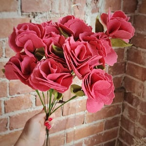 May include: A bouquet of handcrafted felt roses in a vibrant shade of pink. The flowers feature layered, scalloped petals and are attached to thin, dark green wire stems with small, pointed green felt leaves tucked between the blossoms.