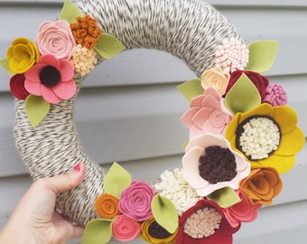 May include: A brown yarn wreath decorated with felt flowers in shades of pink, yellow, orange, and red. The flowers are arranged in a circle around the wreath, creating a festive and colorful display.