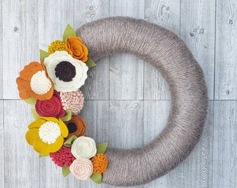 May include: A gray felt wreath decorated with felt flowers in shades of orange, red, white, and yellow. The flowers are arranged in a circular pattern around the wreath.