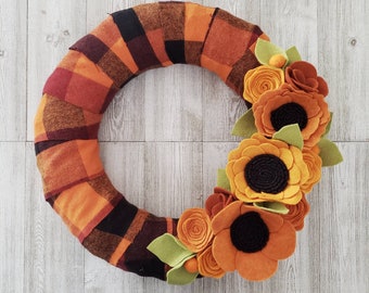 May include: A fall-themed wreath featuring a plaid fabric base in shades of orange, brown, and black. The wreath is adorned with felt sunflowers, orange roses, and green leaves, creating a warm and inviting decorative piece.