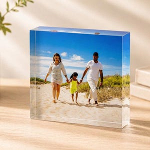 May include: A clear acrylic photo block displays a family walking on a sandy beach under a bright blue sky. The family includes a woman, a man, and a child, all holding hands. The photo block is on a wooden surface.