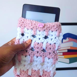 May include: A pink and white crocheted e-reader cover with a pink cat button. The cover is made with a simple stitch pattern and has a slightly textured look.