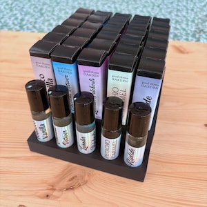 Könnte beinhalten: Ein schwarzer Präsentationsständer mit Reihen kleiner schwarzer Flaschen, die ätherische Öle enthalten. Die Flaschen sind mit Etiketten mit Namen wie "Vanilla", "Vetiver", "Lavender", "Pistachio Caramel" und "Riptide" versehen. Die Flaschen sind in einem Raster auf dem Ständer angeordnet.