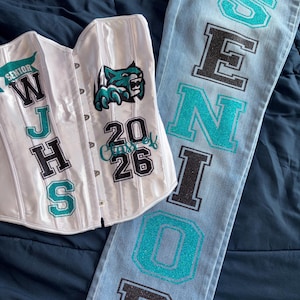 Puede incluir: Un conjunto de corsé blanco y vaqueros azul claro. El corsé presenta el texto "SENIOR W J H S" y un logotipo de pantera verde azulado y negro, con "2026" debajo. Los vaqueros tienen "SENIOR" en letras brillantes verde azulado y negro.