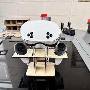 Op de afbeelding: Een witte virtual reality headset met twee zwarte controllers, allemaal gemonteerd op een houten standaard. De headset heeft twee zwarte camera's aan de voorkant.