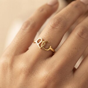 Puede incluir: Un anillo de tono dorado que presenta dos letras interconectadas, una B mayúscula y una C mayúscula, formando un diseño continuo. La banda es delgada y circular, mostrando un acabado metálico pulido que descansa sobre el dedo con sencillez.