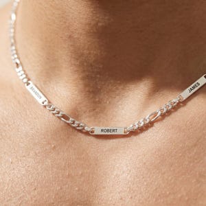 May include: A silver chain necklace with three rectangular nameplates. The nameplates are engraved with the names "BRANDON", "ROBERT", and "JAMES". The necklace has a figaro chain design.