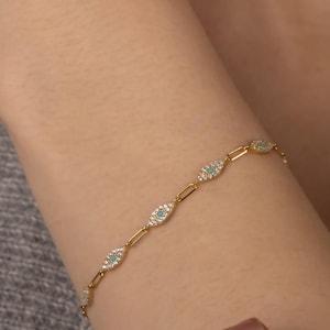 May include: A gold chain bracelet featuring a series of small, oval-shaped charms. Each charm is set with clear cubic zirconia stones and a turquoise blue centre stone, resembling an evil eye.