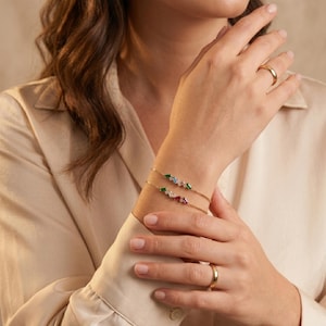 Op de afbeelding: Twee gouden armbanden met kleurrijke edelstenen. Eén armband heeft groene, blauwe en witte stenen, de andere rode, witte en paarse stenen. Er is ook een gouden ring te zien.