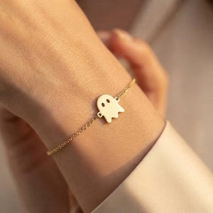Könnte beinhalten: Ein goldfarbenes Armband mit einem kleinen, flachen, geisterförmigen Anhänger, der zwei kreisförmige Aussparungen als Augen aufweist. Der Anhänger ist mittig an einer zarten, dünnen Metallkette befestigt, die das Handgelenk umschließt.