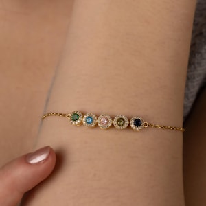 Könnte beinhalten: Ein goldenes Kettenarmband mit sechs kleinen runden Anhängern. Jeder Anhänger hat einen andersfarbigen Edelstein, der von kleineren klaren Edelsteinen umgeben ist.