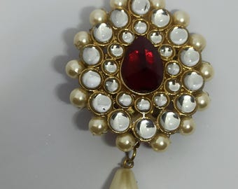 Puede incluir: Broche dorado con una piedra preciosa roja oscura en forma de lágrima en el centro. La broche está rodeada de piedras redondas transparentes y perlas color crema. Un gran colgante en forma de pera, color crema, cuelga en la parte inferior.