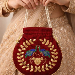 May include: A round, burgundy velvet purse with a drawstring closure and pearl handles. The purse features an embroidered design of two peacocks and gold leaf accents. The purse is approximately 8 inches in diameter.