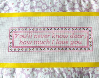 May include: A rectangular needlepoint canvas featuring a pink stitched border with small flower motifs and the text "You'll never know dear, how much I love you" in pink thread. The canvas is framed by a bright yellow tape border on all four sides.