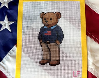 May include: A needlepoint canvas featuring a brown teddy bear wearing a navy blue sweater with a small American flag graphic, a light blue collared shirt underneath, and tan trousers. The canvas has a yellow border and the initials LF in pink.