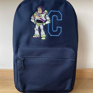 以下が含まれることがあります： ネイビーブルーのバックパック。バズ・ライトイヤーの刺繍と「C」の文字が特徴。フロントにジッパーポケットと調節可能なストラップが付いています。学校や旅行に適した、クラシックなデザインです。