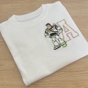 Puede incluir: Camiseta blanca con una imagen bordada de Buzz Lightyear de Toy Story y la letra "A" en una fuente de estilo universitario.