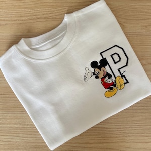 May include: White crew neck sweatshirt with an embroidered Mickey Mouse design and the letter "P". Mickey Mouse is in his classic outfit: red shorts, yellow shoes, and white gloves. The letter "P" is black with a white outline.