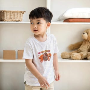 Könnte beinhalten: Ein hellgraues T-Shirt mit dem Text "BE LIKE PUNCH" und einer Cartoon-Grafik eines Affen und eines Faultiers. Das T-Shirt wird von einem kleinen Kind getragen. Das Kind trägt eine khakifarbene Hose.