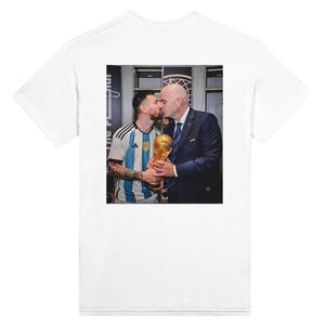 Puede incluir: Una camiseta blanca de manga corta con un estampado gráfico rectangular en la espalda. La imagen muestra a un futbolista con una camiseta de rayas azules y blancas y a un hombre con traje oscuro besando juntos el trofeo dorado de la Copa Mundial.
